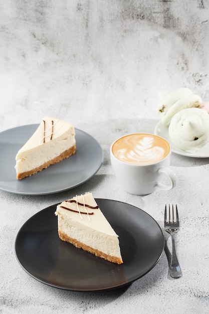 Cheesecake New York or classic cheesecake with cup of coffee on white table. Closeup view. Tasty breakfast. Piece of cake on black plate, white cup on white marble background. Vertical photo.