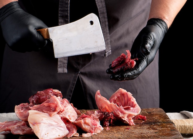 Chef in black latex chunks cuts into pieces raw rabbit meat on a brown wooden board