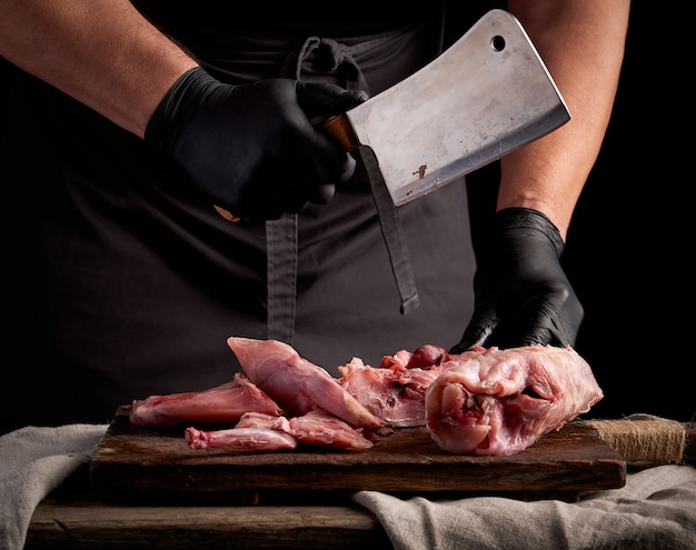 Chef in black latex gloves holds a big knife and cuts into pieces raw rabbit meat on a brown wooden cutting board