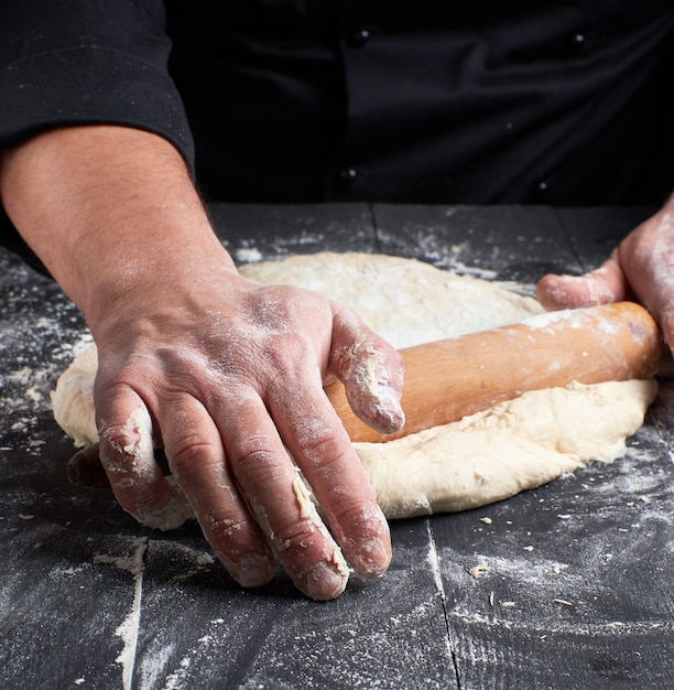 Chef in a black tunic rolls a dough for a round pizza 