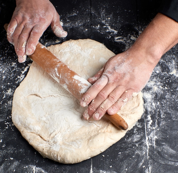 Chef in a black tunic rolls a dough for a round pizza