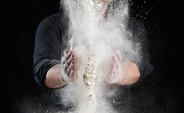 Chef in black uniform sprinkles white wheat flour in different directions, product scatters dust, black background, close up