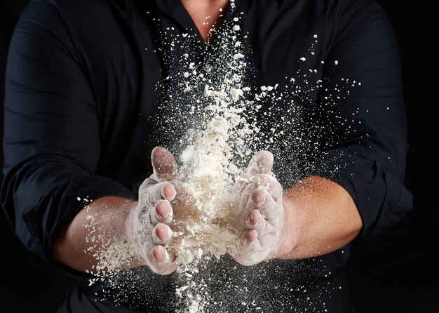 Chef in black uniform sprinkles white wheat flour in different directions, product scatters dust, black background, close up