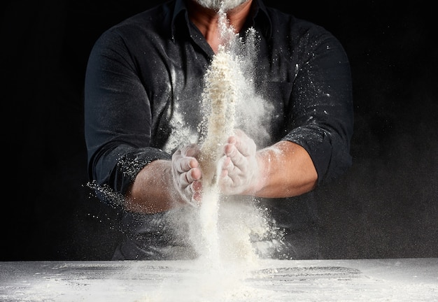 Chef in black uniform sprinkles white wheat flour in different directions, product scatters dust, black space, man sitting at a table