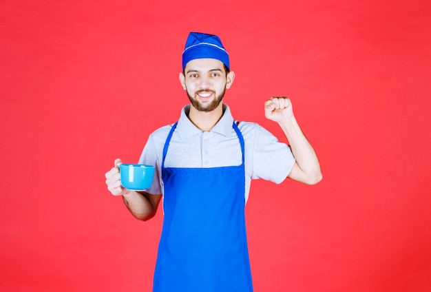 Chef in blue apron holding a blue ceramic cup and showing his fist. 