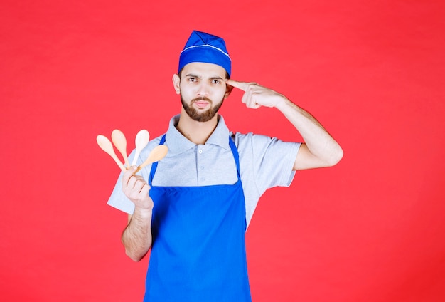 Chef in blue apron holding wooden small spoons and thinking about choice. 