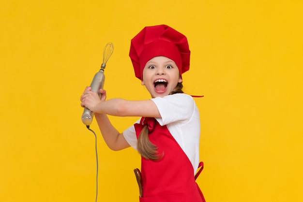 The chef child holds a blender for whipping A happy little girl in a red hat and apron Cooking dishes Yellow isolated background