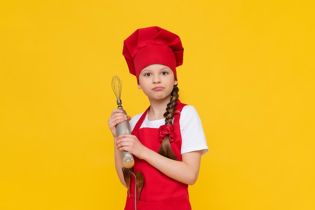 The chef child holds a blender for whipping A thoughtful little girl in a red hat and apron Cooking dishes Yellow isolated background