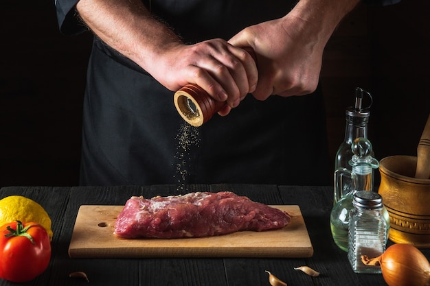 Chef or cook cooking raw meat fillet and adding pepper and chili for marinade on black background. Working environment in the kitchen in restaurant or cafe