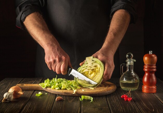 Chef or cook cuts fresh cabbage with knife for salad on vintage kitchen table with fresh vegetables