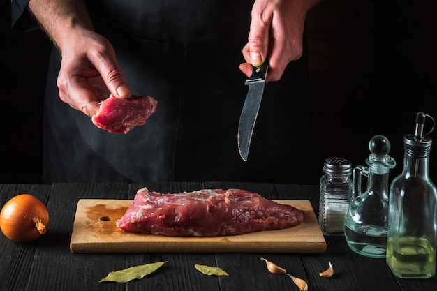 Chef or cook cutting beef meat with knife on kitchen, cooking food. Vegetables and spices on the kitchen table to prepare delicious lunch