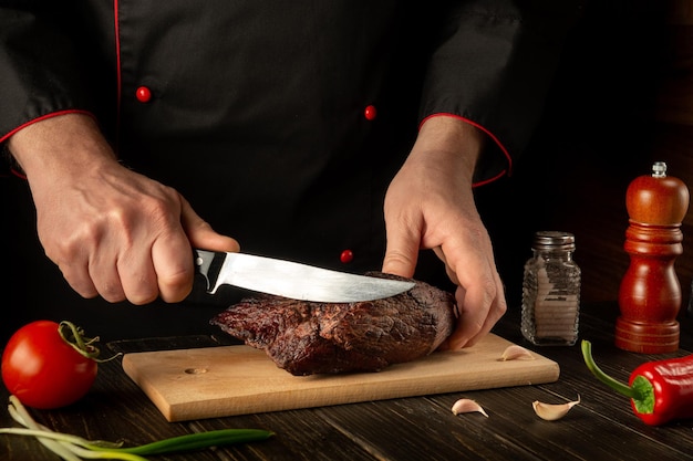 Photo the chef cuts the baked beef steak with a knife on a wooden table the concept of the cooking process