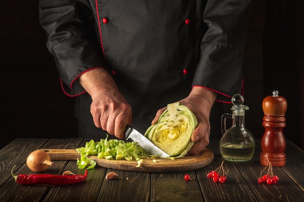 Chef cuts fresh cabbage with knife for salad on a kitchen table with fresh vegetables