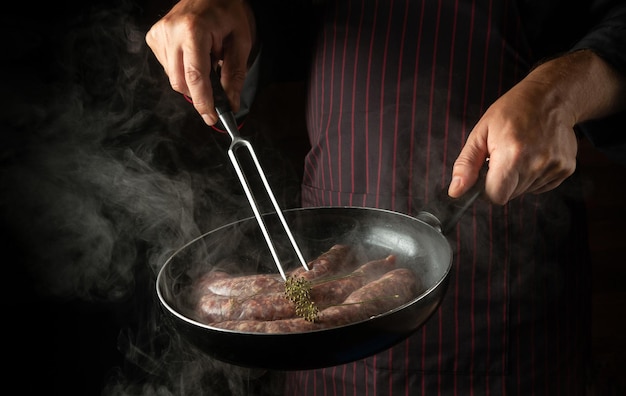 The chef fries sausages for hot dogs in frying pan Closeup of the cook hands with a hot pan and fork