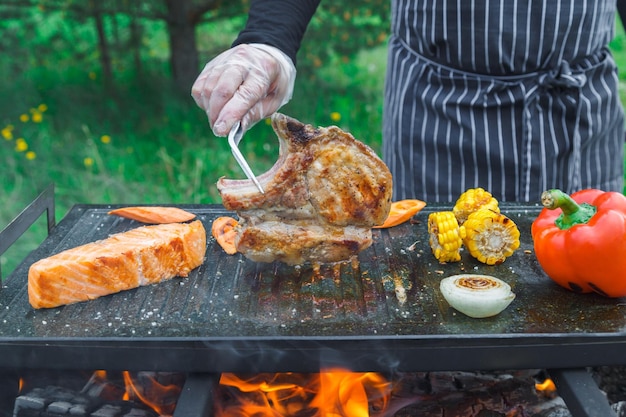 Chef frying meat and vegetables on grill cook in black apron fries red peppers on fire barbecue in o...