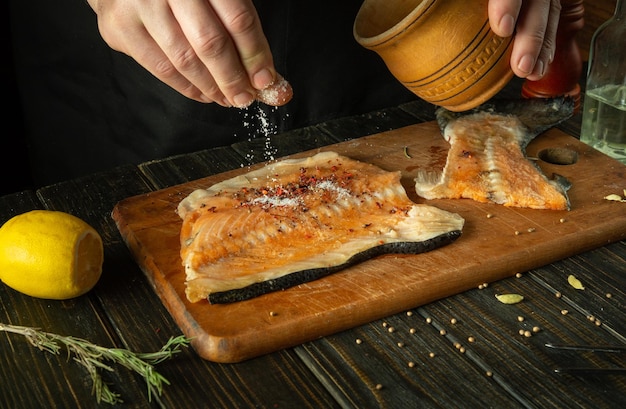 The chef hand adds salt to the fish steak before baking with spices and herbs Concept of cooking pink salmon fish on the kitchen table