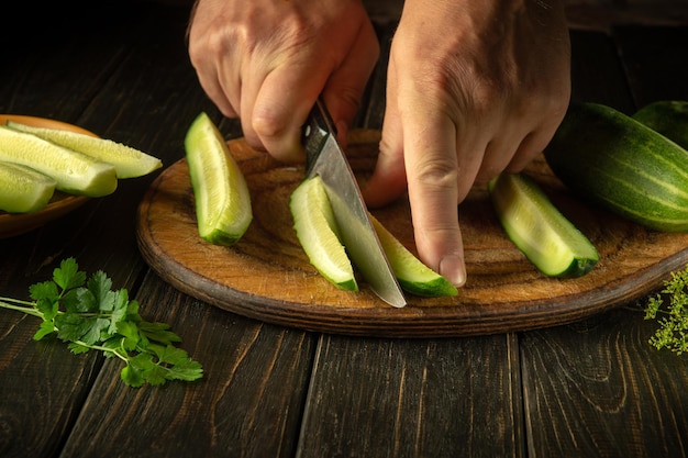 Chef hands with a knife cut fresh green cucumber for salad Working environment in a restaurant kitchen Fresh cucumbers and parsley on the table