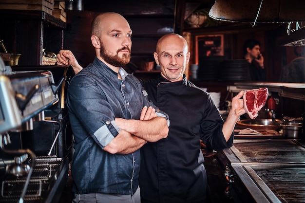 Chef holding a fresh steak and his assistant standing near with his arms crossed in a restaurant kitchen.