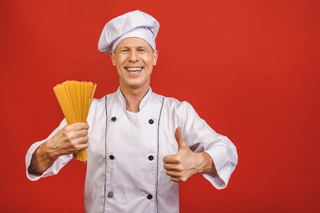 Chef holds bunch of spaghetti in hand. Catering and italian food concept isolated on red background. Cook with satisfied face in white uniform holds dry pasta showing thumbs up.