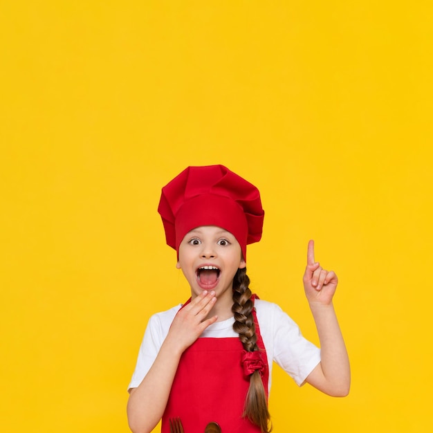 The chef is a little girl in a red hat and apron pointing her finger over her head and your advertisement on a yellow isolated background Copy space