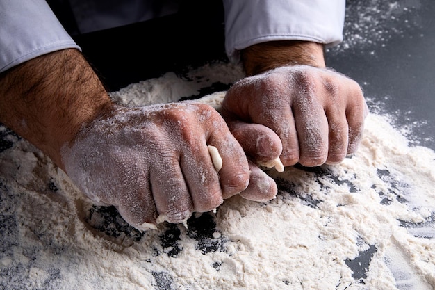 The chef kneads the dough on a dark background, the table is strewn with flour