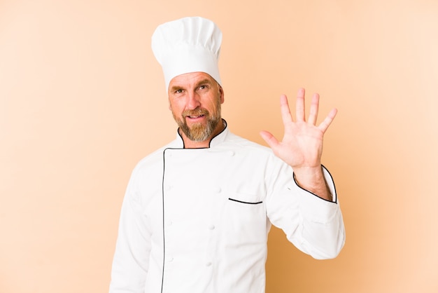 Chef man on beige wall smiling cheerful showing number five with fingers.