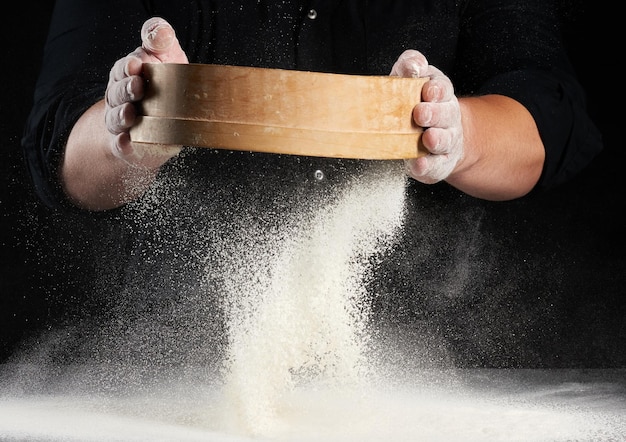 Photo chef a man in a black uniform holds a round wooden sieve in his hands and sifts white wheat flo