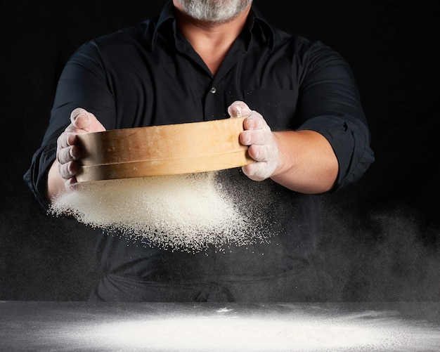 Photo chef a man in a black uniform holds a round wooden sieve in his hands and sifts white wheat flour on a black background, the particles fly in different directions, dusty space