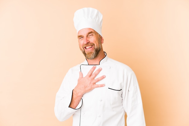 Chef man isolated on beige wall laughs out loudly keeping hand on chest