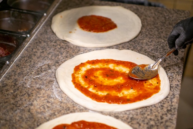 Chef pizza with a metal spoon spreads tomato sauce on the dough in gloves