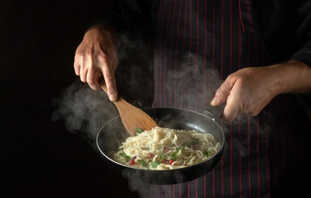 The chef prepares delicious spaghetti with spices and seasonings in a steamed hot pan Space for advertising on a dark background Menu for a restaurant or hotel