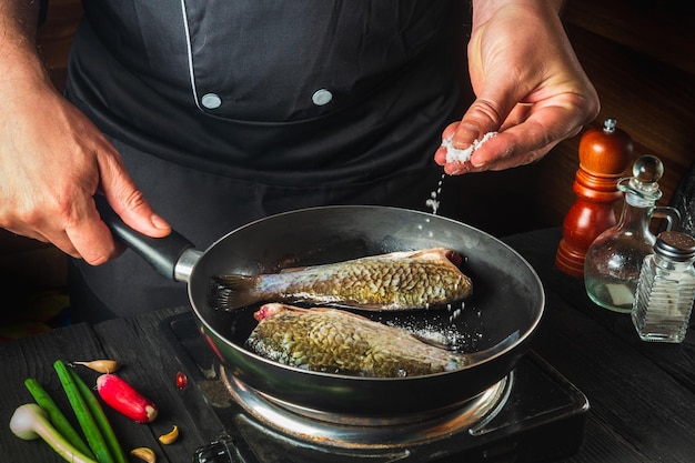 Chef prepares fresh fish in pan sprinkling salt with the ingredients. Preparing to cook fish food. Working environment in the restaurant kitchen.