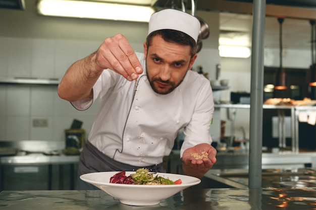 Chef sprinkles salt to salad of fresh vegetables in kitchen of restaurant Tasty and healthy food