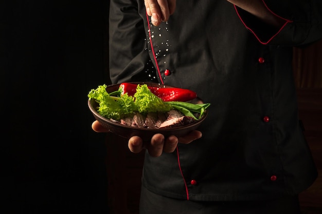 The chef sprinkles salt on a sliced steak with beef and vegetables in a plate