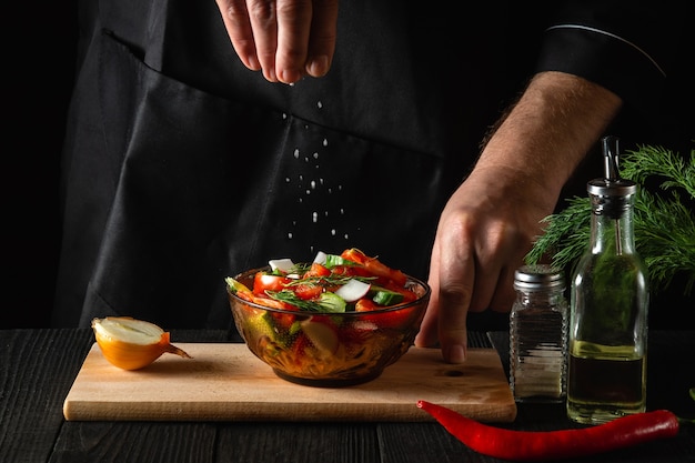 Chef sprinkles salted fresh vegetable salad in a plate on a wooden table