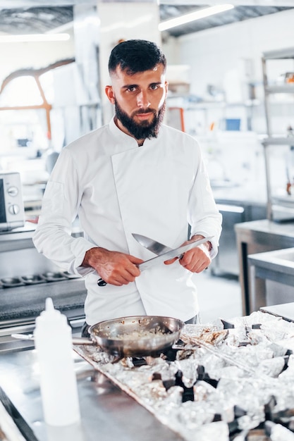 Chef in white uniform standing at kitchen Holding knives in hands