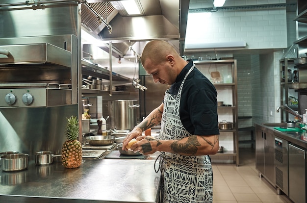 chef with several tattoos on his arms peeling a grapefruit while standing in a professional kitchen