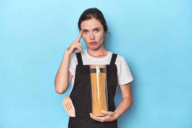 Chef woman with pasta jar on blue pointing temple with finger thinking focused on a task