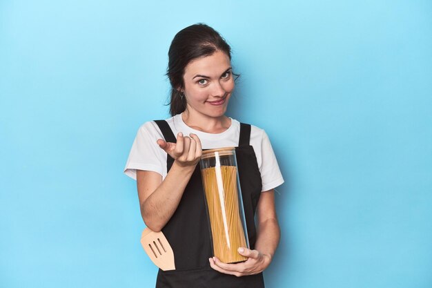 Chef woman with pasta jar on blue pointing with finger at you as if inviting come closer