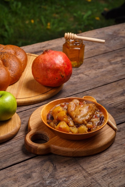 Chelnt with chicken in a wooden plate with a wooden spoon on the festive table for Rosh Hashanah next to apples challah and pomegranate