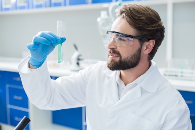 Chemical experiment. Nice positive serious man wearing protective glasses and holding a test tube while doing a chemical experiment