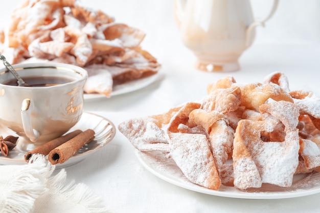 Chenchi carnival sweets on a white plate. Sprinkled with powdered sugar. Nearby is a cup of tea decorated with cinnamon. White background.