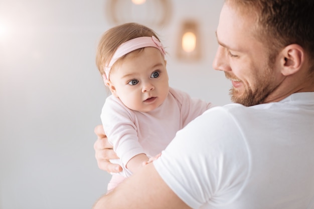 Cherishing every moment. Cheerful smiling bearded father standing at home and hugging the toddler girl while expressing tender emotions