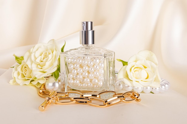 A chic perfume bottle is filled with pearls against the background of beige waves of satin roses a gold necklace Front view Creative composition