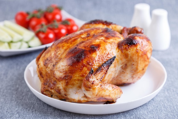 Chicken baked whole to a delicious crust A traditional dish In the background are cucumbers and cherry tomatoes on a white plate Closeup selective focus gray background