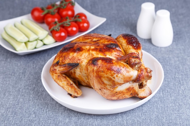 Chicken baked whole to a delicious crust A traditional dish In the background are cucumbers and cherry tomatoes on a white plate Closeup selective focus gray background