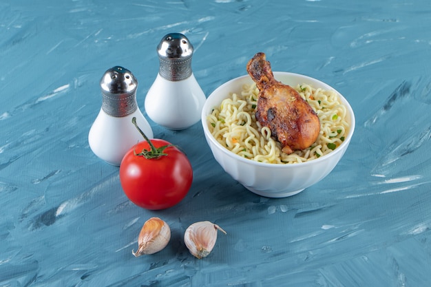 Chicken drumsticks and noodle in a bowl next to salt, tomatoes and garlic, on the marble surface.