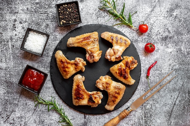 Chicken legs on a black plate with spices and sauces on a gray background.