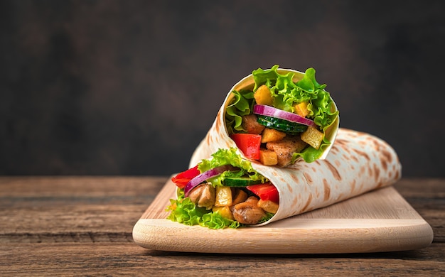 Chicken meat, French fries, vegetables and salad are wrapped in pita bread on a brown wall. Traditional shawarma. Side view, copy space.