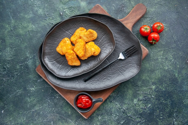 Chicken nuggets on a black plate and fork on wooden board tomatoes ketchup on dark surface